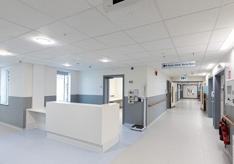Hospital corridor with a white and grey color scheme. Features include a reception desk with a digital display, red fire extinguishers, light blue and grey tiled flooring, and various signs above doors. The ceiling has recessed lighting