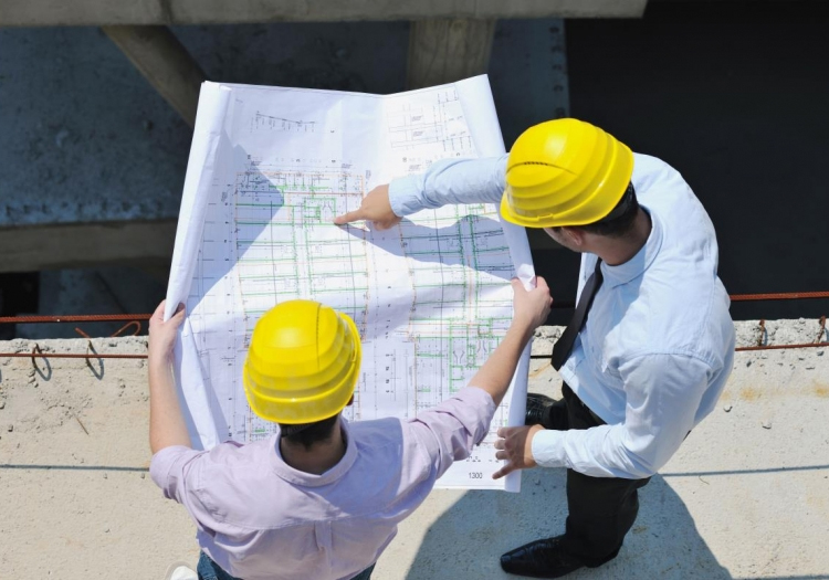 Two individuals in yellow hard hats and white shirts, viewed from above, examining a large sheet of paper with architectural plans or engineering drawings. The focus on the detailed plans indicates they are likely professionals discussing a project.
