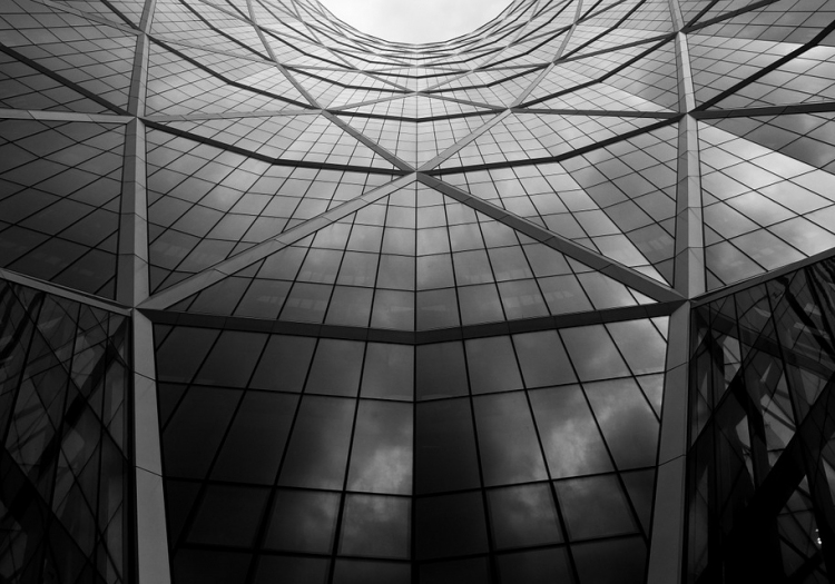 Black and white photograph of a modern building with a geometric design, featuring curved and straight lines. The glass facade reflects the sky and clouds, creating depth and contrast."
