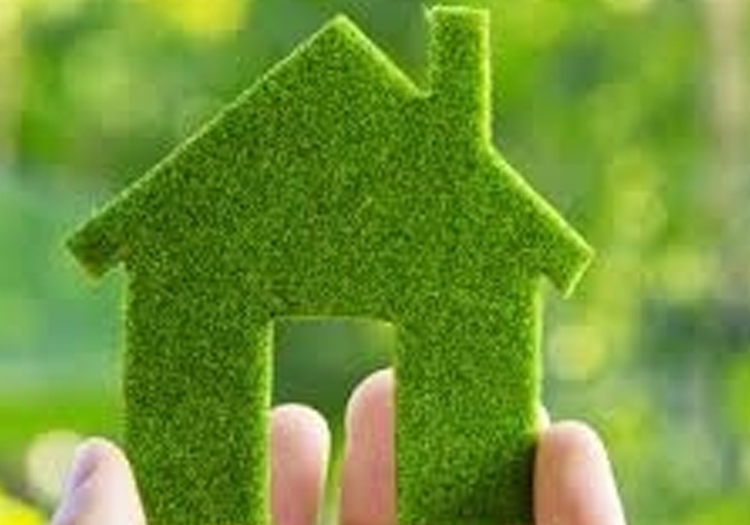 Pair of hands holding a small house-shaped object covered in greenery, resembling grass or moss. The blurred background suggests an outdoor setting, likely a garden or park. The image represents eco-friendly living, sustainable housing, or integrating nature into daily life.