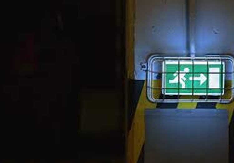 Illuminated green exit sign with a white arrow pointing right, visible through a dark window or opening, indicating emergency exit direction