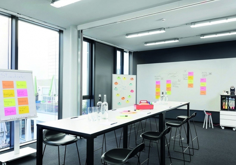 Modern meeting room with natural light from a large window. Features a long black table with chairs, water bottles, glasses, notebooks, and pens. Includes a whiteboard with markers, a shelf with office supplies and sticky notes, and a back wall covered in organized sticky notes.