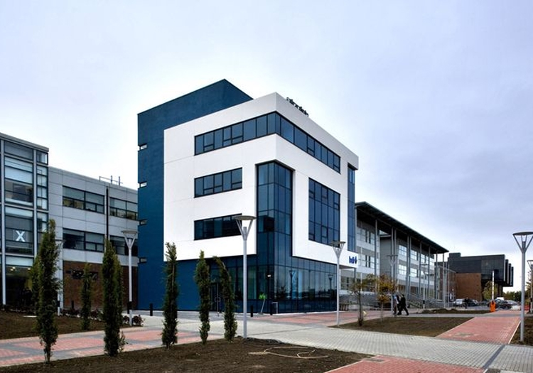 Modern building with blue and white exterior walls, part of a larger complex with connected glass-facade structures. The architecture features geometric shapes and clean lines for a contemporary look. In front, a paved area with street lamps and young trees at intervals suggests an educational or corporate campus.
