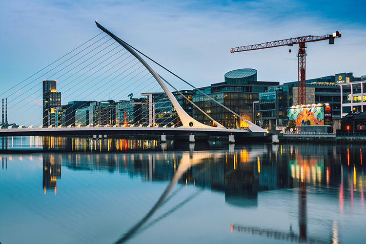 Modern cityscape at twilight with a cable-stayed bridge, reflecting lights on the water, and buildings in the background.