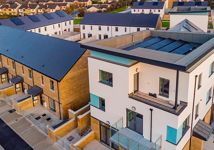 Aerial view of a modern residential area with houses featuring flat and sloped roofs, built from brick, wood, and render. Each house has a balcony or patio, illuminated by warm sunrise or sunset light, emphasizing the contemporary design and layout.