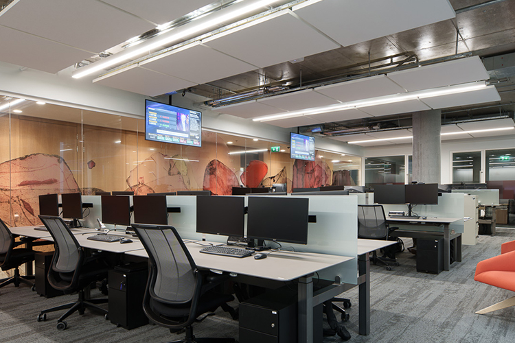 Modern open-plan office with workstations, dual monitors, ergonomic chairs, and orange accent chair. Features concrete column, glass partitions with red artwork, and wall-mounted TV displaying financial data