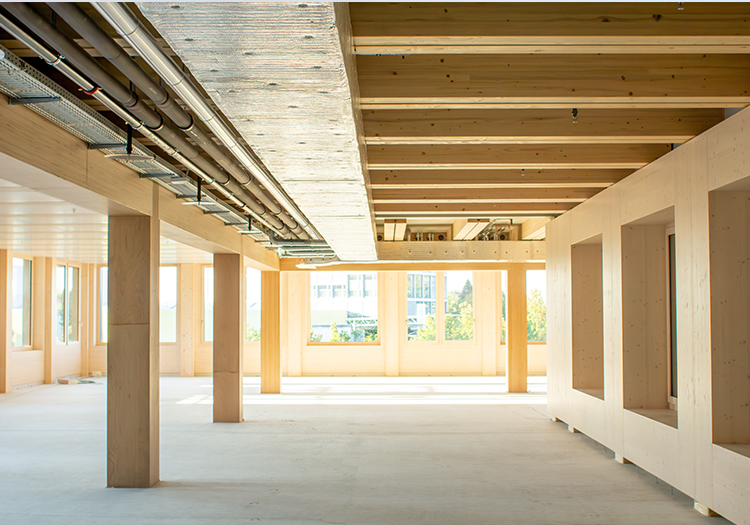 Interior of a commercial building with exposed cross-laminated timber structure, visible service ducts, and large windows allowing natural light.