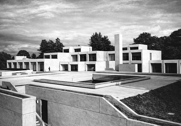 Modern concrete building with geometric forms arranged around a stepped outdoor courtyard, photographed in black and white