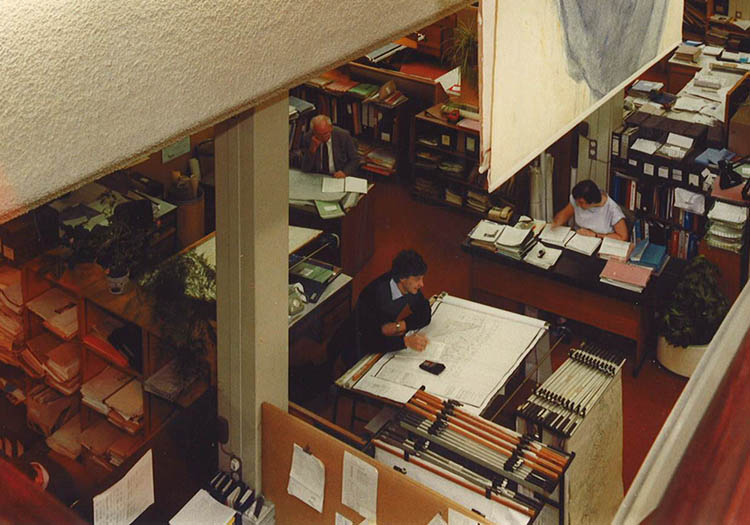 Engineering team reviewing technical drawings in a studio workspace with drafting tables and design documents.