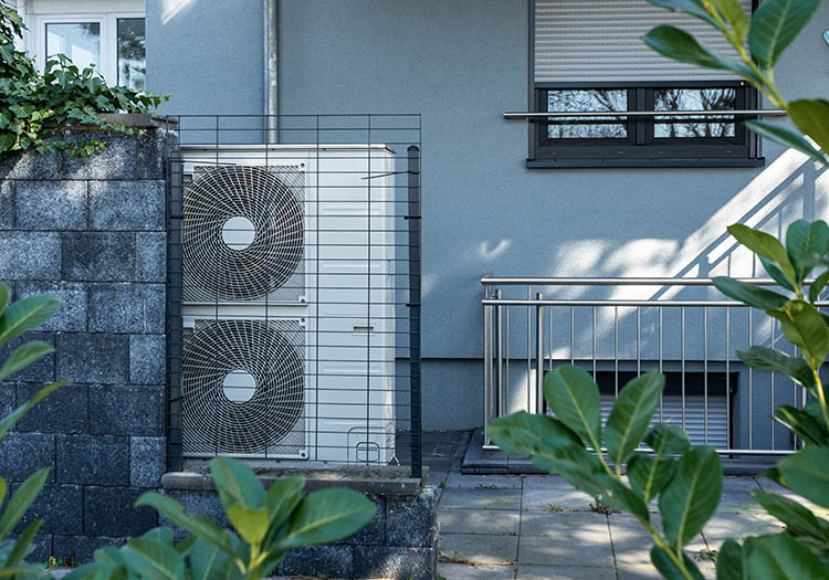 Outdoor monobloc air source heat pump unit installed beside a residential building and protected by a metal enclosure